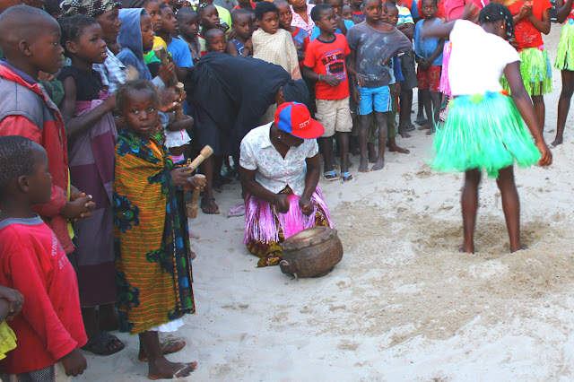 Na piasku plaży nad jeziorem w Bilen stoi półkolem grupa afrykańskich dzieci. Dzieci ubrane są kolorowo. Kilkoro z nich trzyma w dłoniach proste, drewniane kołatki do wybijania rytmu. Inne klaszczą w dłonie. Siedząca na piasku kobieta gra na bębnie.Obok tańczy dziewczynka w jaskrawozielonej spódnicy.