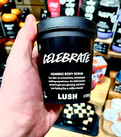 A white hand holding a black cylindrical large tub filled with yellow scrub with a black label that says celebrate foaming body scrub in white font on a bright background