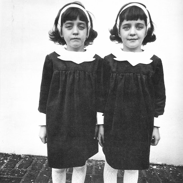 Foto em preto e branco, mostrando duas meninas gêmeas, de vestidos iguais e uma tiara na cabeça. A da direita sorri, a da esquerda está com o rosto fechado