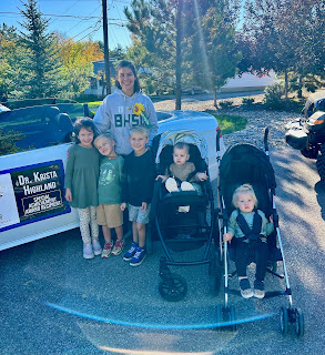 Outdoor photo featuring a smiling person in a grey hoodie standing next to a white convertible car with banners reading 'Dr. Krista Highland' and 'Special Achievement Award Recipient'. Four young children are standing beside them, and two toddlers are seated in a double stroller.