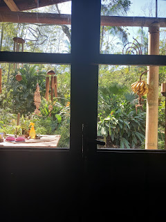 Blick aus Fenstertür, im Hintergrund Bäume, Bambus, blauer Himmel, vorne Windspiele unter Hausdach