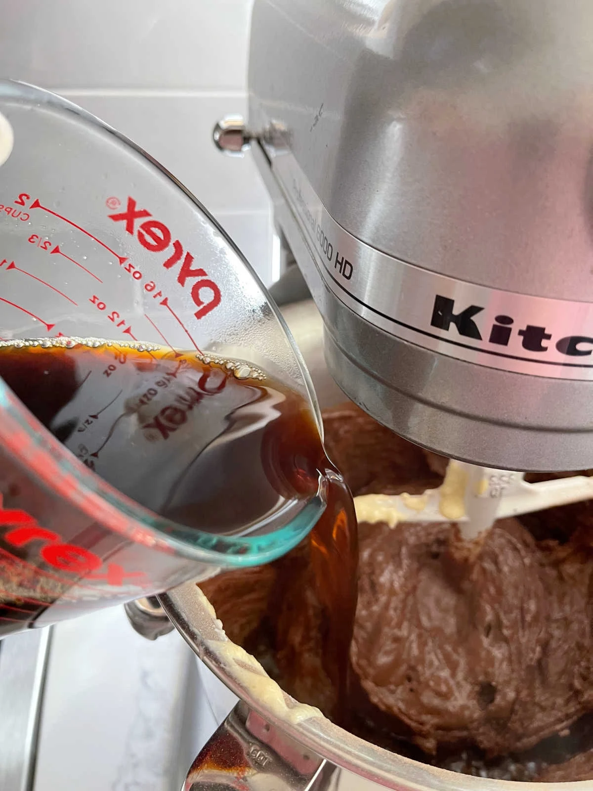 Hot Coffee being poured into Chocolate Cake batter in a stainless steel mixing bowl from a glass measuring cup.