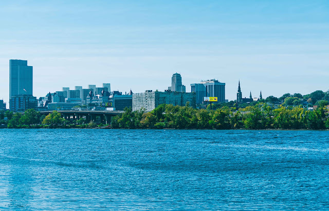 A serene waterfront view of a Albany skyline on a clear day. The cityscape includes a mix of modern high-rises and the famous Central Warehouse, with church spires rising elegantly against the backdrop of the blue sky. Lush greenery lines the near side of the water, providing a natural contrast to the urban structures. Gentle ripples on the surface of the water reflect the bright sunlight.