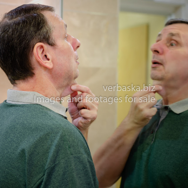 A mature man lost large amounts of weight. He touches his saggy skin in the throat area and looks in the mirror so surprised. 