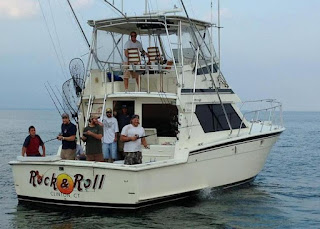 picture of men standing on the stern of a a deep sea fishing boat, there are many fishing poles on the white boat. The name of the boat is Rock and roll. It is floating in the open ocean on slightly choppy waves. That are appearing to have a lot of fun.