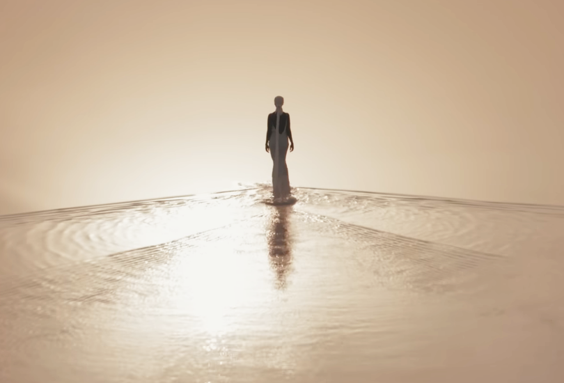 A shot of Kelela in her music video for “Washed Away”, walking through shallow waters at sunset in a white dress and a head scarf.