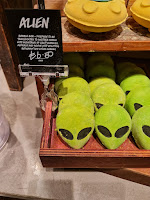 A photo of a square light brown box containing a bunch of oval green alien head shaped bubble bars with black eyes and a black smile next to a black rectangular card that says alien bubble bar in white font  on a light brown rectangular table on a bright background