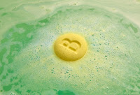 A macaron shaped bath bomb , one side is blue  and purple with a b engraved into it and one side is yellow that has lush engraved into it slowly dissolving into a tub of water on a bright background
