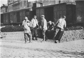 Black and white photo of a brick-building factory with containers in front of it. In front of the container, one the right side stands a stack of cylinder shaped military munition. In front of the container and to the left of the stack stand six men wearing factory boots, thick winter jackets and berets.