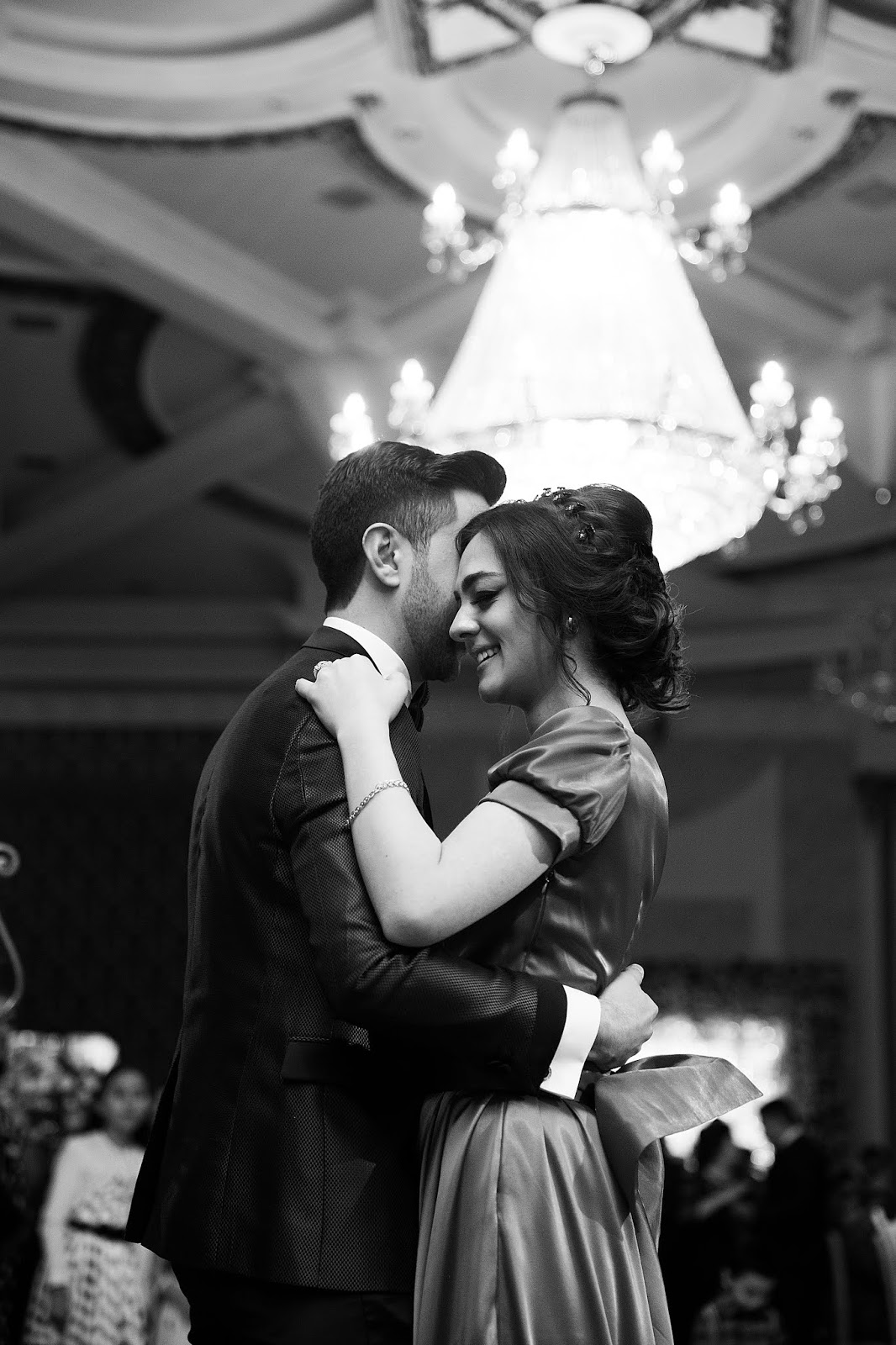 A black and white photo of a a main in a suit and a woman in a silky looking dress with a bow on the back, danicng - the man on the left, the woman on the right, the right side of the top of her head against his right cheek - there are people in the background, a chandelier above, behind, and to the right of them