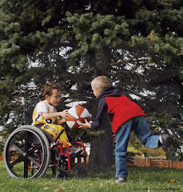 Two boys playing in a yard, with a  red and white basketball. One is in a wheelchair, handing the ball to the other, who is running to grab it