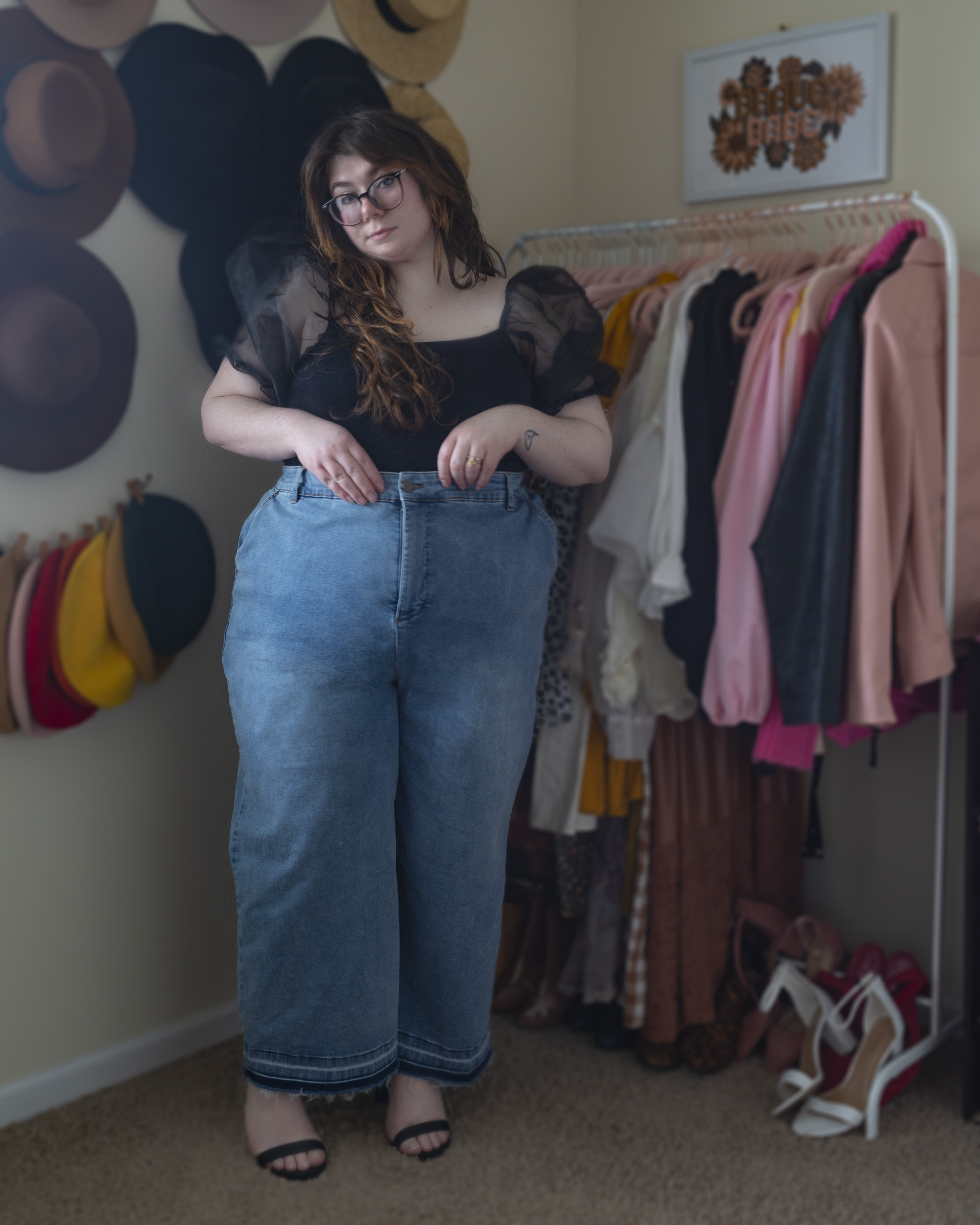 An outfit consisting of a black short organza sheer puffy sleeve with ribbed knit bodice blouse, tucked into light wash wide leg high waist jeans and black ankle strap sandal heels.