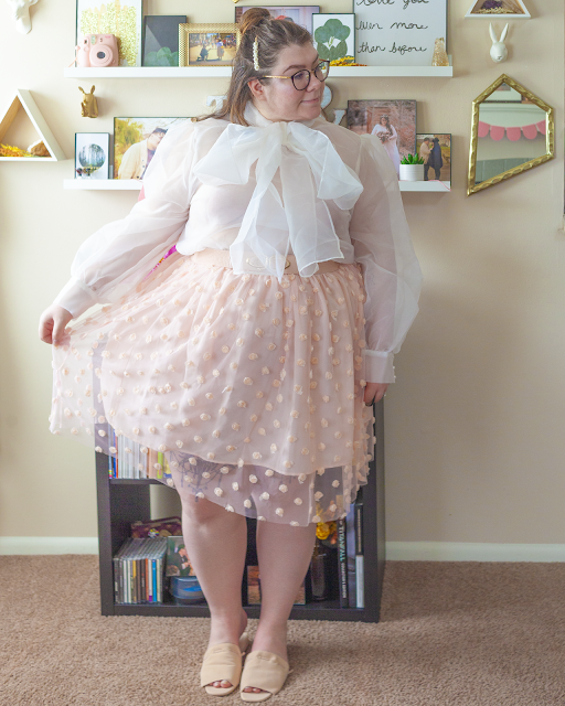 An outfit consisting of a sheer long sleeve blouse with an attached bow tucked into a pastel pink midi skirt with a sheer overlay with rosettes attached to it and pastel pink slide sandals.