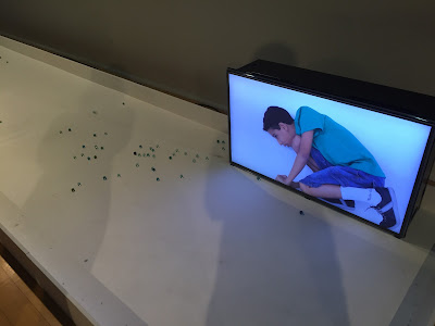 TV com imagem de um menino jogando bola de gude, TV num espaço cheio de bolinhas de gude