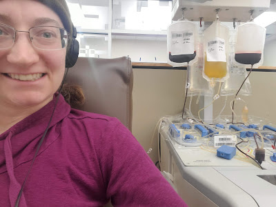 A woman wearing a hot pink shirt and a grey and brown hat with headphones and glasses donates platelets. To the side is the machine with a bag of plasma and two of blood visible in addition to the tubing.