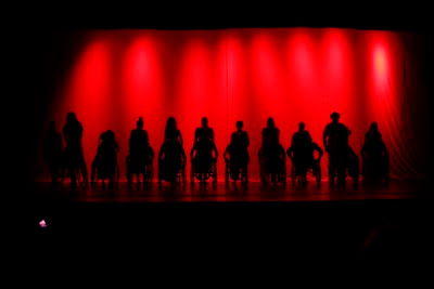 Elenco do musical Filhos do Brasil em silhueta no palco sobre um fundo iluminado de vermelho; cadeirantes lado a lado e atrás de cada cadeira um ator andante.