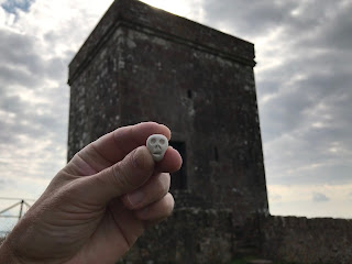 A photo of a small, ceramic skull (Skulferatu 89) being held up with Repentance Tower in the background.  Photograph by Kevin Nosferatu for the Skulferatu Project.