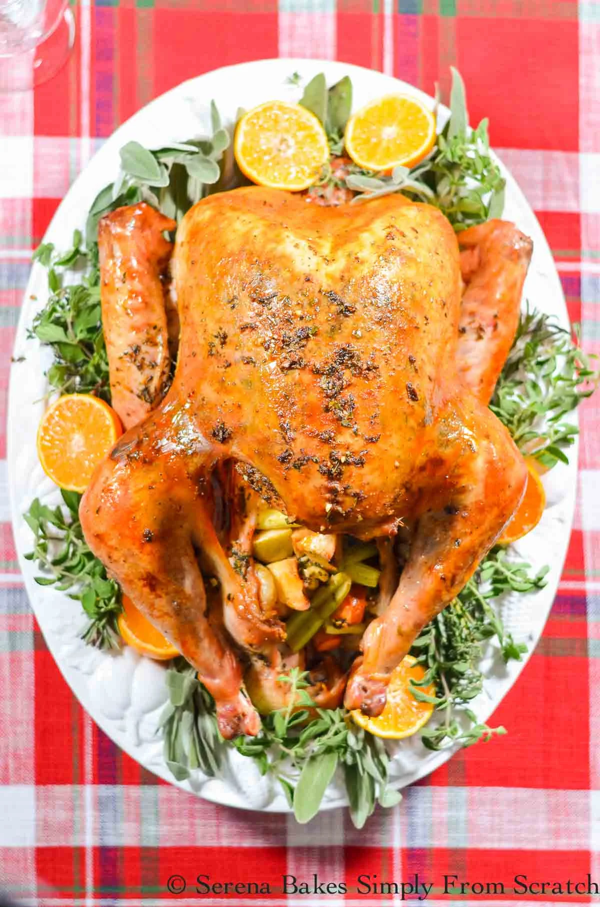Golden Brown Cajun Roasted Cheesecloth Turkey on a white serving platter garnished with fresh herbs and sliced oranges on a red plaid table cloth.