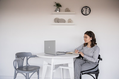 A young woman sits at a kitchen table taking notes in a notebook while looking at a computer screen.