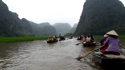 The Tam Coc (“three caves”) portion is a three-hour excursion by small boat along the Ngô Đồng river,  beginning at the village of Van Lam and proceeding through a scenic landscape dominated by rice fields and karst towers. The route includes floating through three natural caves (Hang Ca, Hang Hai, and Hang Ba), the largest of which is 125m long with its ceiling about 2m high above the water. The boats are typically rowed by one or two local women who also sell embroidered goods.