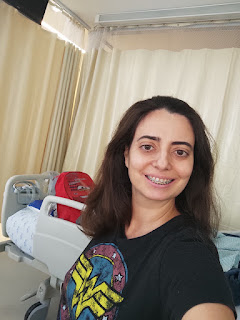 Sabrina usando camiseta preta da mulher maravilha, em pé, em frente a uma cama hospitalar, com a mochila vermelha do carros em cima da cama.