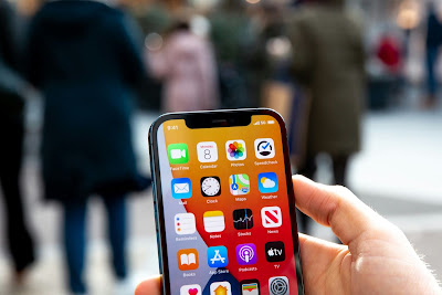 Una mano sosteniendo un iPhone, con la pantalla mostrando las Apps del iPhone de menú principal con un fondo anaranjado. Detrás se muestran varias personas caminando en la calle.