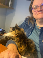 A woman sitting at a desk with a cat in her lap. The cat is draped over her arm.