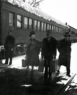 A couple stands holding their luggage as they prepare to travel on a train; the train operator and another man stand behind them in the wintry scene. 1942