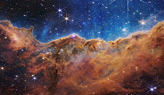 Cosmic Cliffs in the Carina Nebula (NIRCam Image) - The image is divided horizontally by an undulating line between a cloudscape forming a nebula along the bottom portion and a comparatively clear upper portion. Speckled across both portions is a starfield, showing innumerable stars of many sizes. The smallest of these are small, distant, and faint points of light. The largest of these appear larger, closer, brighter, and more fully resolved with 8-point diffraction spikes. The upper portion of the image is blueish, and has wispy translucent cloud-like streaks rising from the nebula below. The orangish cloudy formation in the bottom half varies in density and ranges from translucent to opaque. The stars vary in color, the majority of which have a blue or orange hue. The cloud-like structure of the nebula contains ridges, peaks, and valleys – an appearance very similar to a mountain range. Three long diffraction spikes from the top right edge of the image suggest the presence of a large star just out of view.