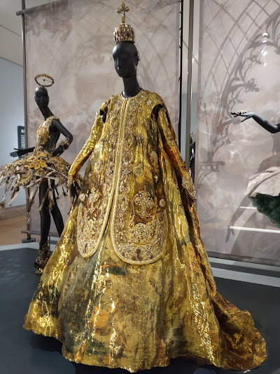 Photo of a gold floor-length dress, flaring out from the shoulders, in a heavily brocaded fabric, with a sort of vest in similar fabric. The mannequin has a ball-shaped gold hat with a cross on it.