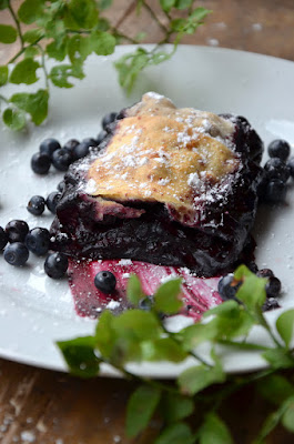 Fertig gebackener Waldheidelbeer-Strudel auf einem weißen Teller angerichtet, daneben liegen Waldheidelbeer-Blätter.