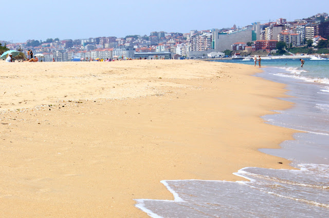 Plaża na piaszczystej mierzeji, zwanej El Puntal, z widokiem na leżące na przeciwległym brzegu Santander. Jasny piasek, fale wdzierające się na plażę, kilka osób siedzących pod parasolami włożonymi w piasek, kilka osób spacerujących nad brzegiem, a w tle posadowiione na wzgórzu, ciasno zabudowane kamienicami i blokami miasto.