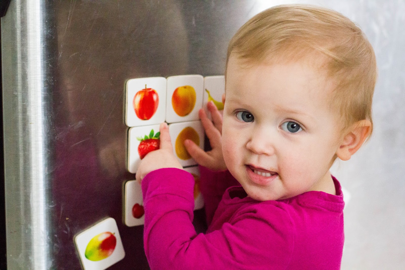 Young toddler stands in front of refrigerator pointing to various wooden magnets with pictures of fruit.