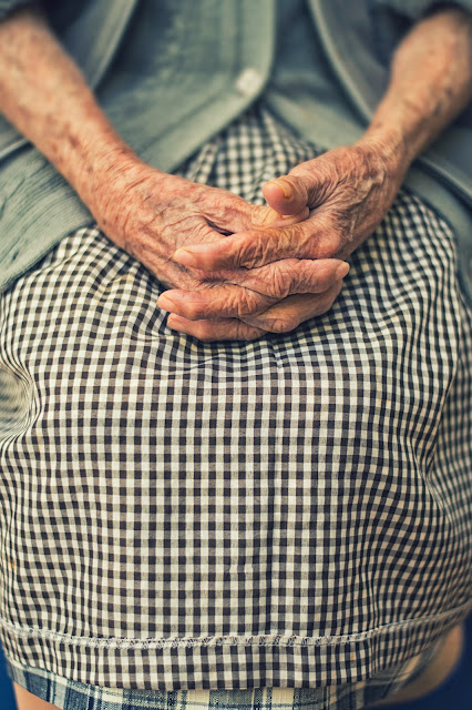 Elderly woman wearing gingham skirt with wrinkled hands folded on lap:Photo by Danie Franco on Unsplash