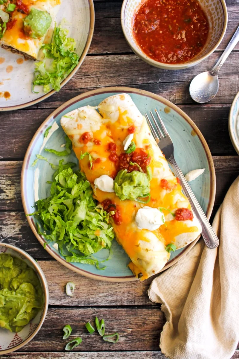 Top view of two burritos on a blue plate topped with a spoonful of greek yogurt, salsa, and guacamole on top with a side of shredded lettuce.  Plate is on a dark wood background.