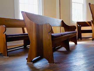Photograph of a wooden bench in a Quaker meeting room. Other benches are visible in the background, and the sun shines through windows further in the background.