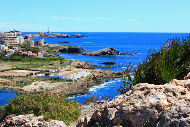 widok z klifów na Cabo de Palos. Urozmaicane niewielkimi, kamiestymi wysepkami wybrzeże, z półwyspem pełnym niewielkich domów, ponad dachy których wystaje smukła sylwetka latarni morskiej. Niebo i morze mają nasycony, ostro błękitny kolor. 