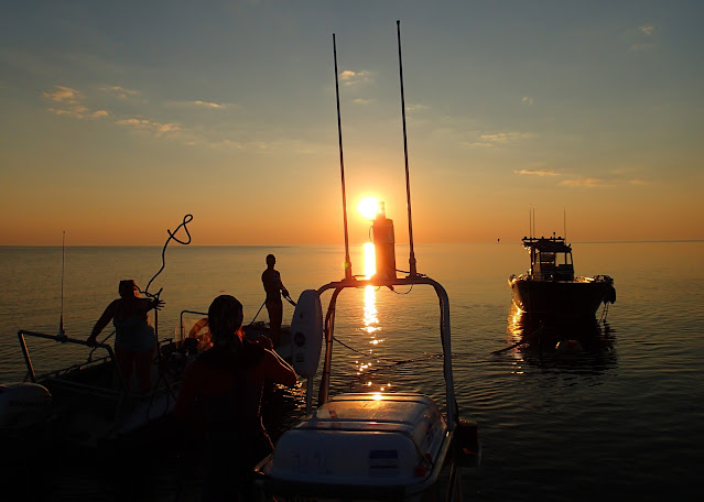 Auringonlasku merellä, rannalla pari venettä ja ihmisiä silhuetteina