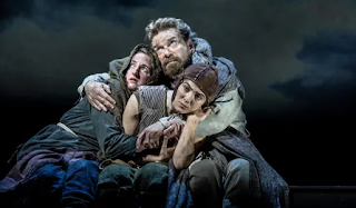 Kenneth Branagh, as King Lear, is hugging towards him two other characters. He's got bushy hair and a beard, and wears furs and earth-tone robes.