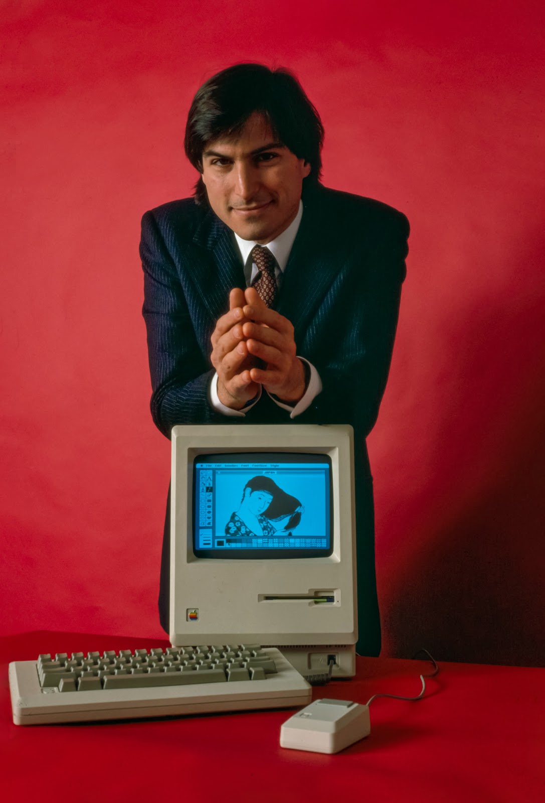Macintoshの筐体の上に両肘を付いて微笑むスティーブ・ジョブズ