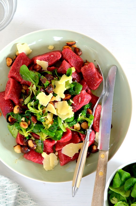 Rote Gnocchi aus roten Rüben sind auf einem Teller platziert, darauf Vogerlsalat, Haselnüsse und würziger Parmesan.