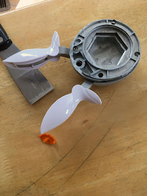 A photo from above of a metal hexagonal paper punch with two long white plastic handles on a light wood table. A small orange plastic lozenge in sitting on the table, and a narrow metal drywall scraper has been set down near the partly-disassembled punch.