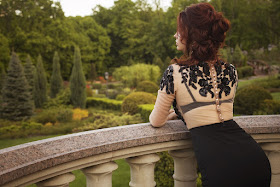 A dark-haired woman in a black lace dress leanimg on a balcony looking out at the backyard.