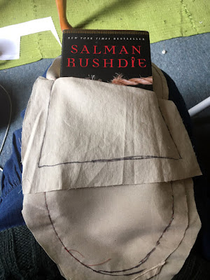 A beige roughly-oval pocket, with seam lines marked in black sharpie and large seam allowances, sitting on the photographer's lap, with the top half of Salman Rushdie's book 'Shalimar the Clown' showing over the front edge.