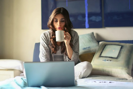 woman drinking from a white mug with a laptop on a bed