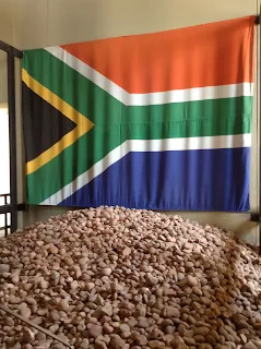 South African flag hanging on a white wall. In front of the flag is a tall pile of small brown rocks