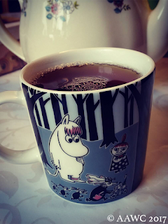 A mug of tea in front of a teapot. The mug has a Moomin character on it.