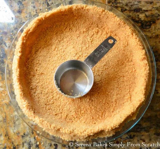 Graham Cracker crumbs mixture being pressed into a pie plate with a silver measuring cup.