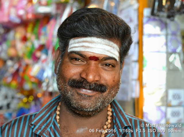 Matt Hahnewald Photography; Facing the World; closeup; street portrait; headshot; flash fired; Asia; South Asia; India; Tamil Nadu; Tiruchirappalli; Nikon D3100; Nikkor AF-S 50mm f/1.8G; travel; travel destination; souvenir salesman; Hindu; Hinduism; photography; colour; portraiture; person; people; eyes; black hair; face; Srirangam; Sri Jambukeshwara Temple; eye contact; vibhuti; white ash on forehead; smiling; tilaka; souvenir shop; Tamil man; tripundra; Shiva devotee