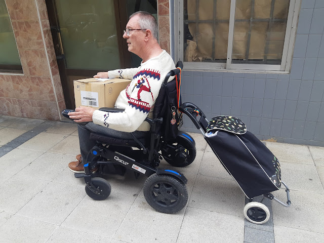 Un hombre sentado en una silla de ruedas lleva una caja con libros y un carrito a remolque